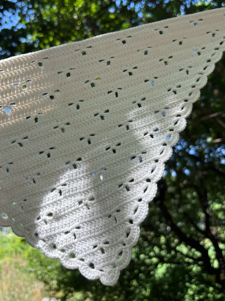 Close-up of a white crocheted fabric held up outdoors, with sunlight casting a cat's shadow onto the fabric and green leaves visible in the blurred backgroundโperfect inspiration for your next summer crochet pattern or when searching for a crochet bandana pattern free.