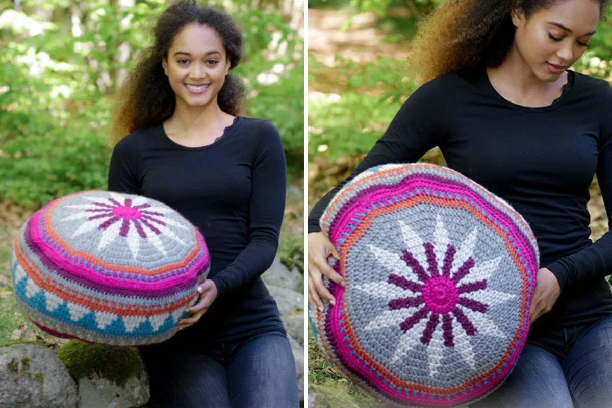 Woman holding a round, colorful crocheted Marrakesh Pouf cushion with pink, gray, and blue patterns, outdoors in a green, natural setting.