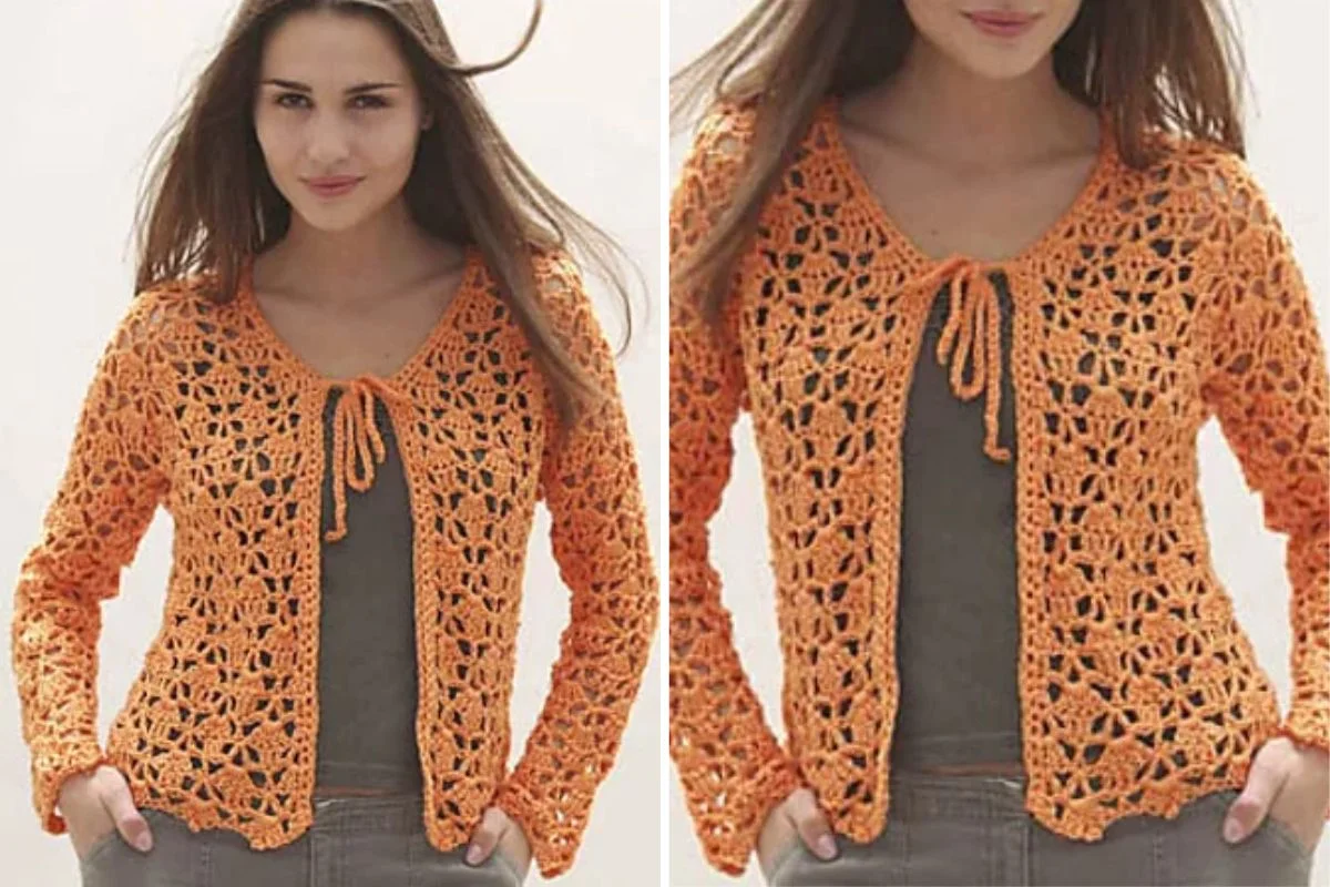 Woman wearing a long-sleeve, orange crocheted cardigan with a tie at the neck over a dark top and grey pants, standing against a light background.