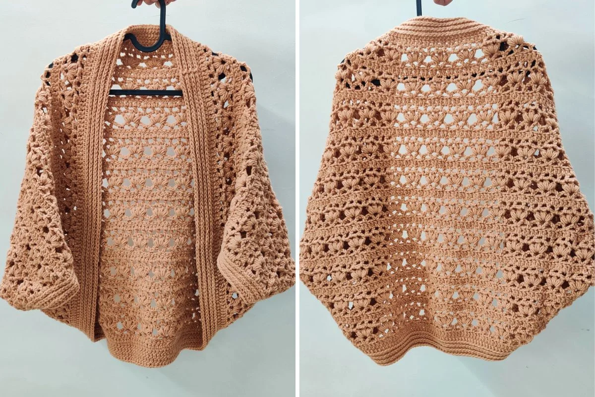 A crocheted tan cardigan is shown from the front and back on a hanger, featuring an openwork, lacy pattern and wide sleeves.
