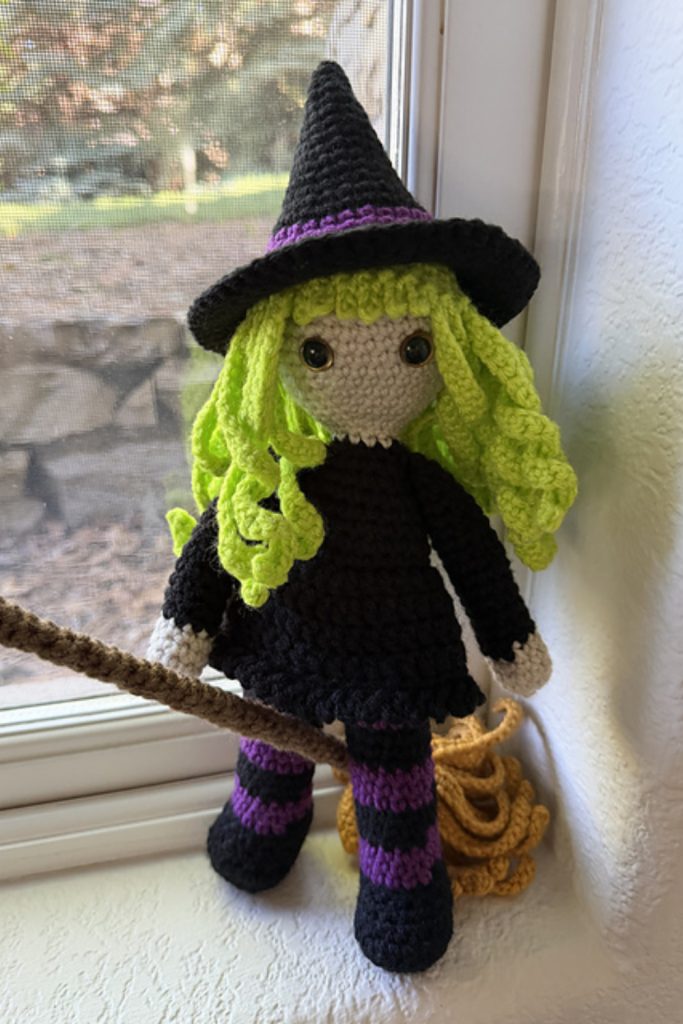 A crocheted doll dressed as a witch with a black hat, green hair, black dress, and striped purple-and-black stockings sits on a window ledge holding a broomstick.