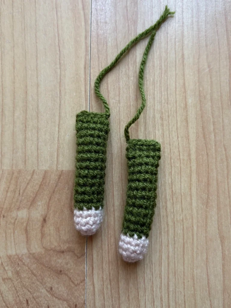Two crocheted green and beige objects with strings attached, lying parallel on a wooden surface.