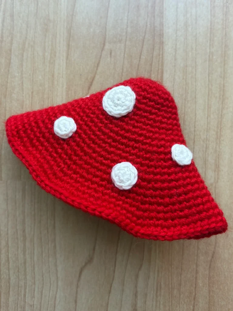 Red crocheted object shaped like a mushroom cap with four white crocheted spots, reminiscent of a Toadstool Gnome, placed on a light wooden surface.