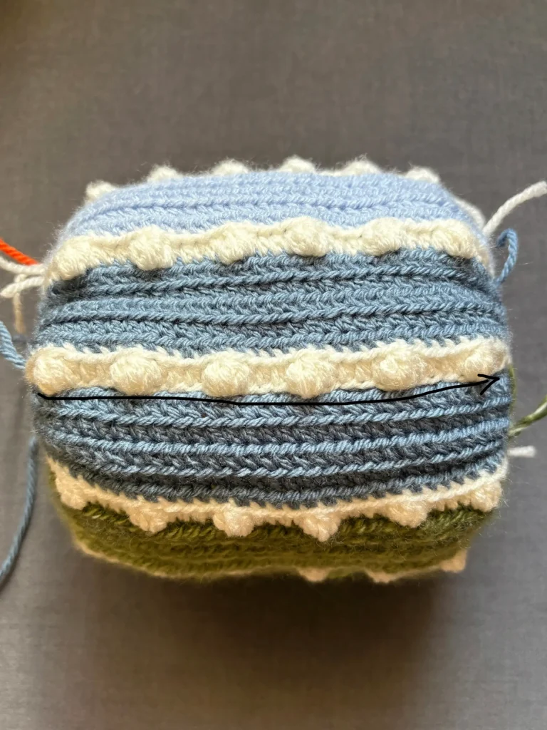 A crocheted rectangular object with blue, green, and white stripes, featuring textured rows and loose yarn ends, sits on a gray surfaceโan inviting project for those searching for inspiration beyond a pumpkin crochet pattern or the whimsical Toadstool Gnome.