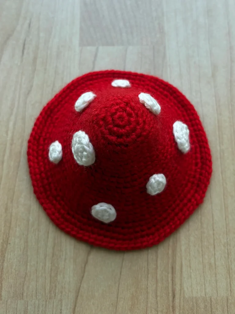 A red crocheted object with white spots, shaped like a mushroom cap, is placed on a light wooden surface. If you enjoy whimsical crochet designs, you might also love exploring a free crochet pumpkin pattern to complement this adorable piece.