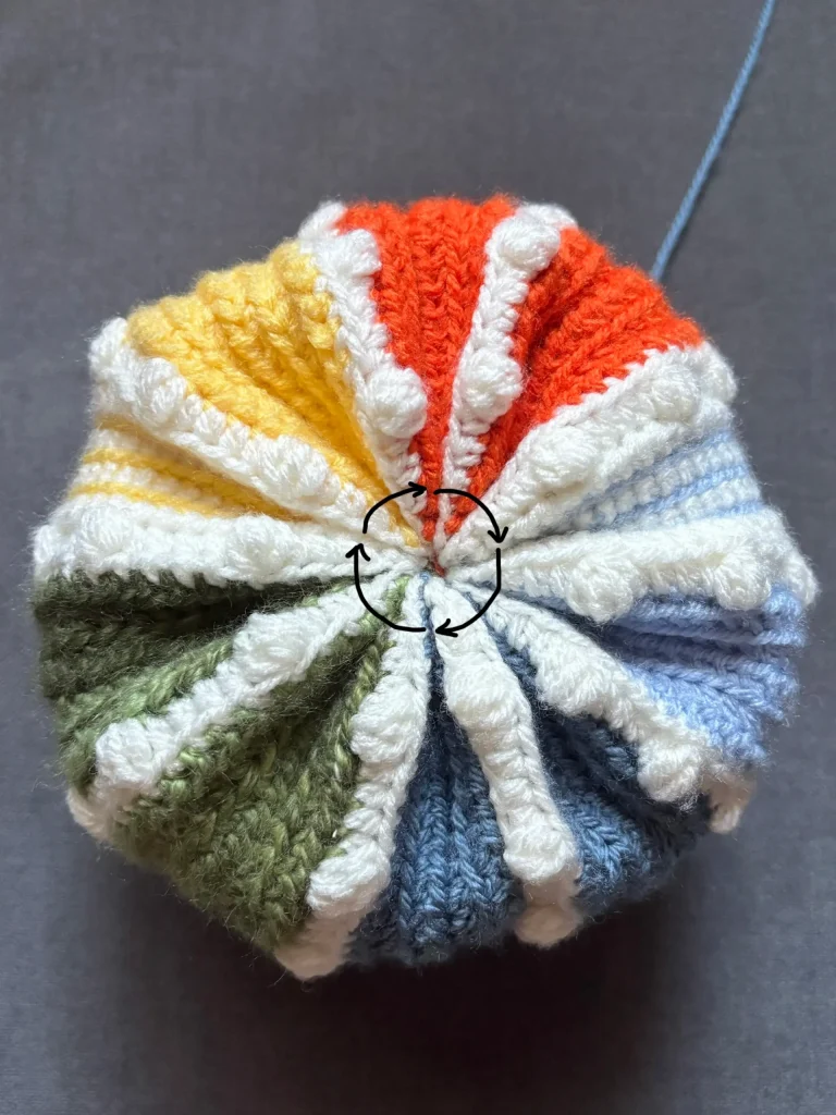 A crochet, round object with colorful sections and a central point where the yarn is gathered, reminiscent of a pumpkin crochet pattern, is shown against a gray background.