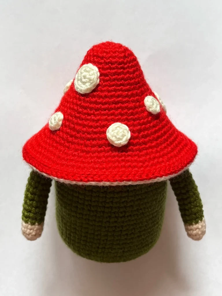 A crocheted doll inspired by the Toadstool Gnome, featuring a red, white-spotted mushroom cap, green body, and pink-tipped arms, is shown against a plain white background.