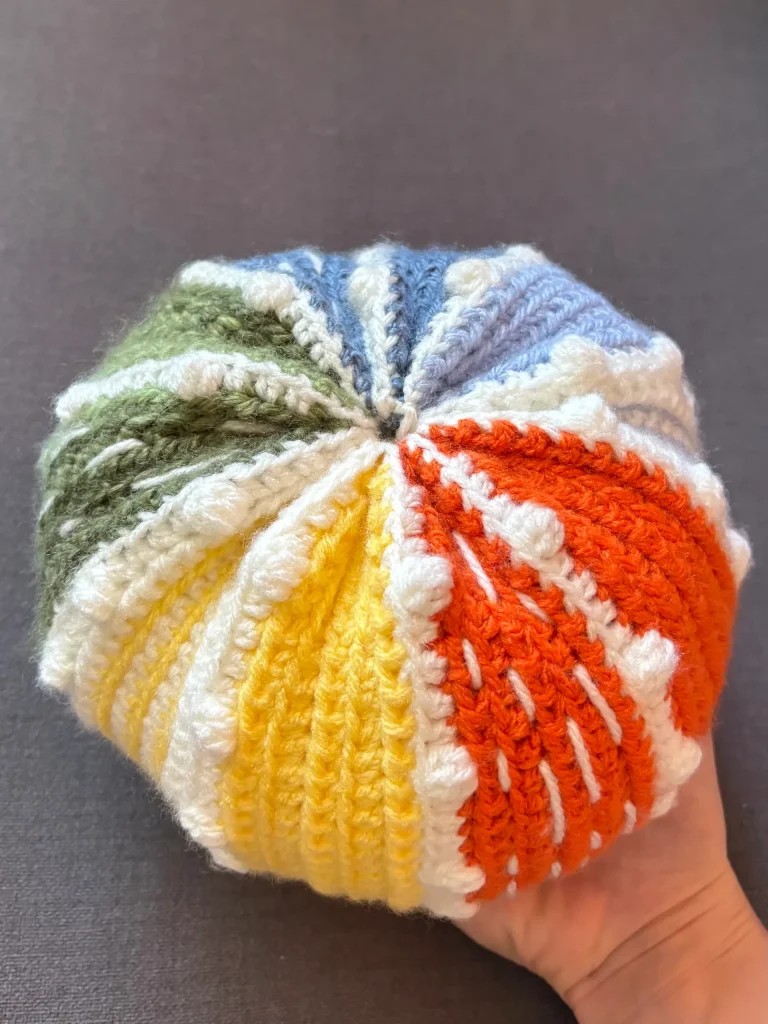 A hand holds a crochet round object featuring alternating segments of orange, yellow, green, blue, and white yarn on a plain gray background, resembling a playful twist on a pumpkin crochet pattern.