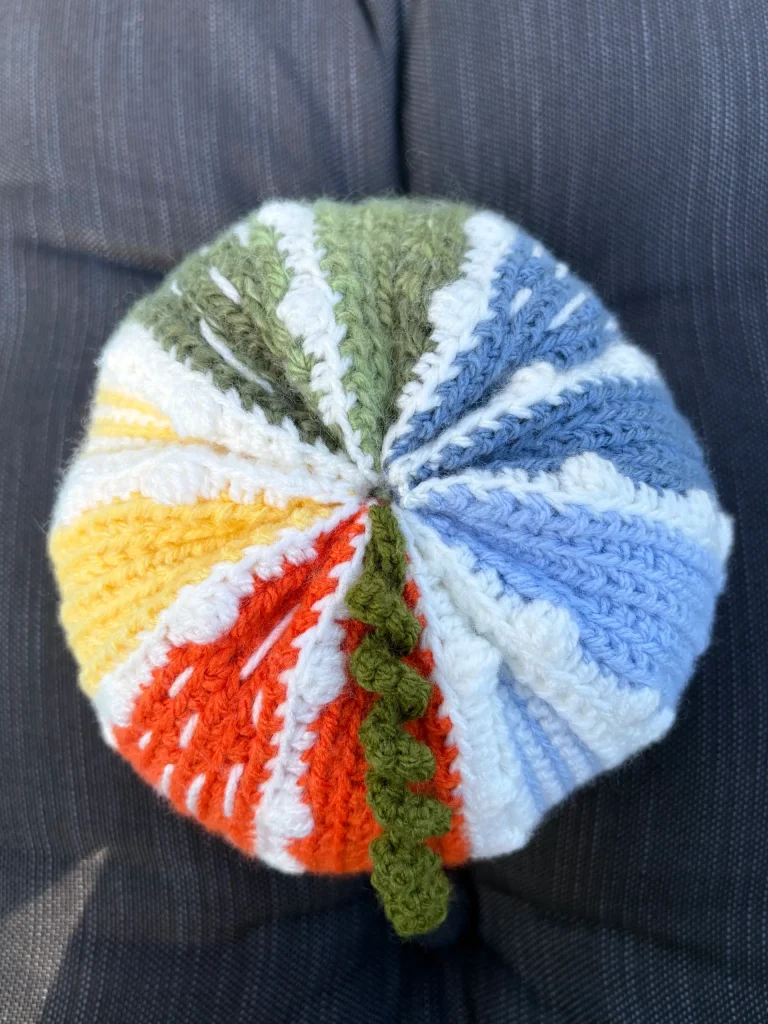 A knitted or crocheted hat with orange, yellow, green, blue, and white sectionsโreminiscent of a cozy pumpkin crochet patternโsits on a dark, textured fabric background.