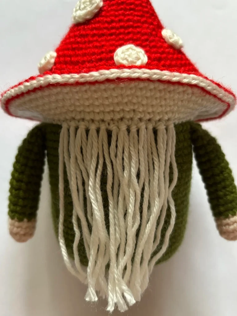 A charming Toadstool Gnome figure is featured with a red mushroom cap, white yarn beard, green body, and beige hands, expertly crocheted in delightful detail. Perfect for fans of whimsical decor or those searching for inspiration beyond the classic free crochet pumpkin pattern, this gnome brings a touch of woodland magic to any display.