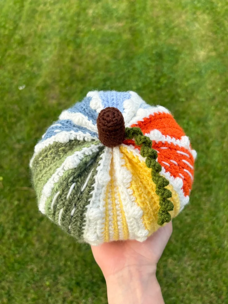 A hand holds a vibrant knitted pumpkin crafted using a free crochet pumpkin pattern, with sections in green, yellow, orange, white, and blue yarn and a brown stem, set against a grassy background.