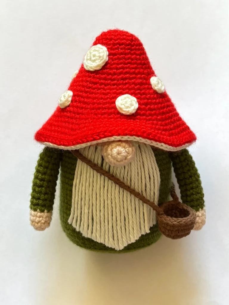 Toadstool Gnome crocheted doll with a red mushroom cap hat, white beard, green body, and a brown satchel, on a white backgroundโa charming handmade figure thatโs perfect for fans of whimsical designs and those searching for inspiration beyond the classic crochet pumpkin pattern.