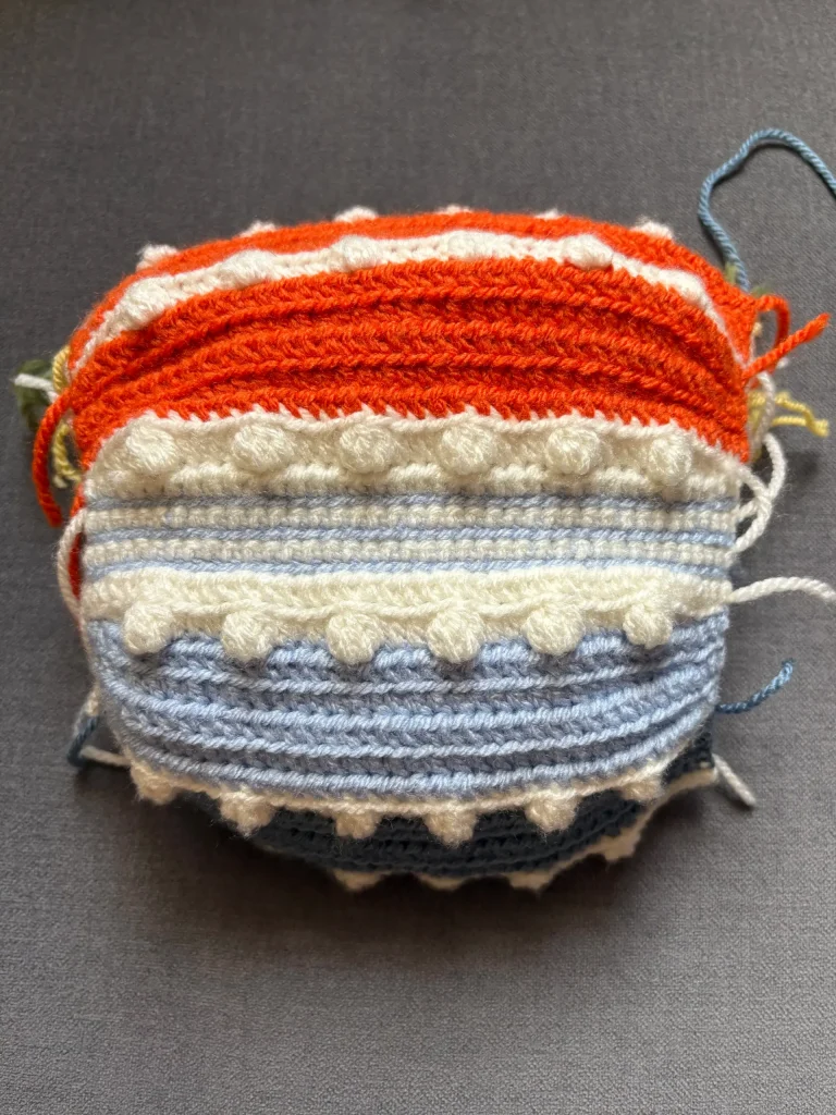 A swatch of textured crochet fabric in orange, white, and blue yarn, featuring horizontal stripes and raised bobble stitches reminiscent of designs seen in a free crochet pumpkin pattern, lying on a gray surface.