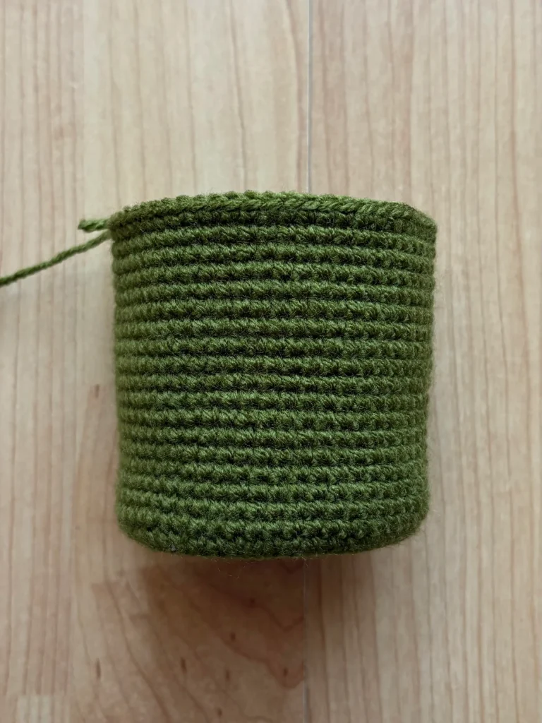 A cylindrical object crocheted with green yarn, reminiscent of a pumpkin crochet pattern, sits on a light wood surface.