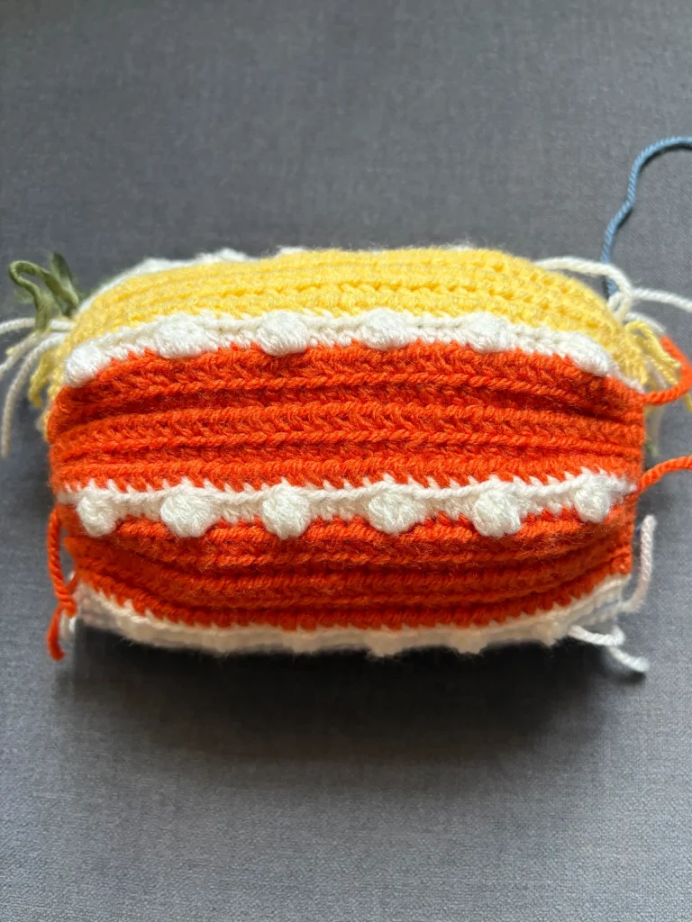 A rectangular piece of crocheted fabric in yellow, orange, and white yarn with textured and bobble stitchesโreminiscent of designs from a free crochet pumpkin patternโis resting on a gray surface.