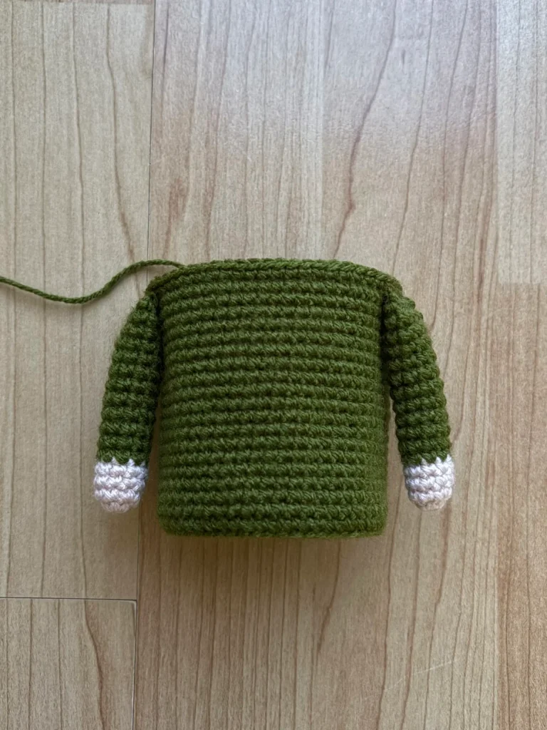 A crocheted green torso with long sleeves and white cuffs lies on a wooden surface; no head or legs are attached, making it a charming work-in-progress that could easily complement your favorite free crochet pumpkin pattern.
