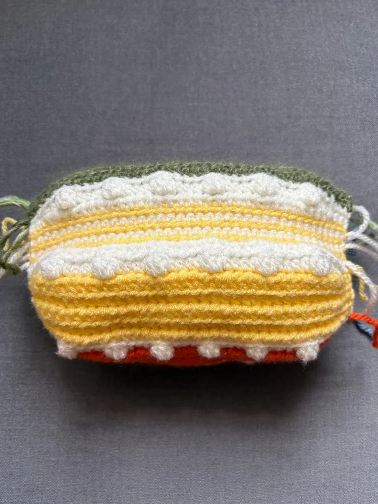 A rectangular crocheted pouch with horizontal stripes in green, white, yellow, and orange, featuring textured patterns and loose yarn ends on a gray backgroundโperfect for storing your next free crochet pumpkin pattern or keeping your favorite pumpkin crochet pattern handy.