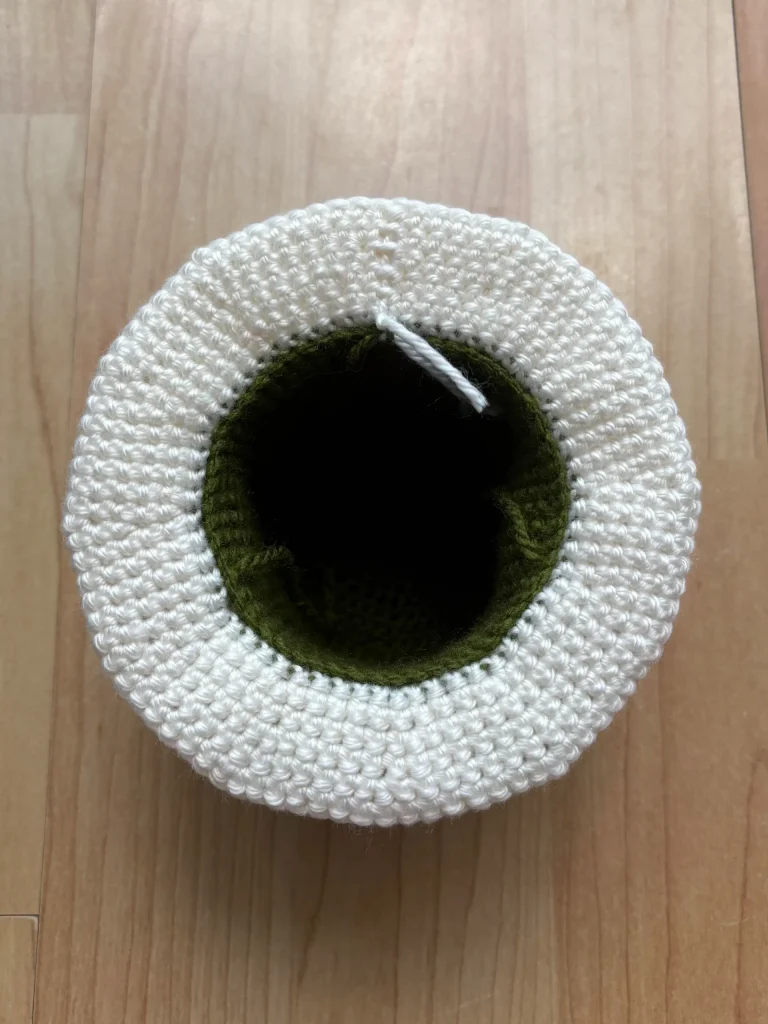 A crocheted object with a white outer layer and green inner section, seen from above, placed on a light wood surfaceโperfect inspiration if youโre searching for a unique pumpkin crochet pattern.