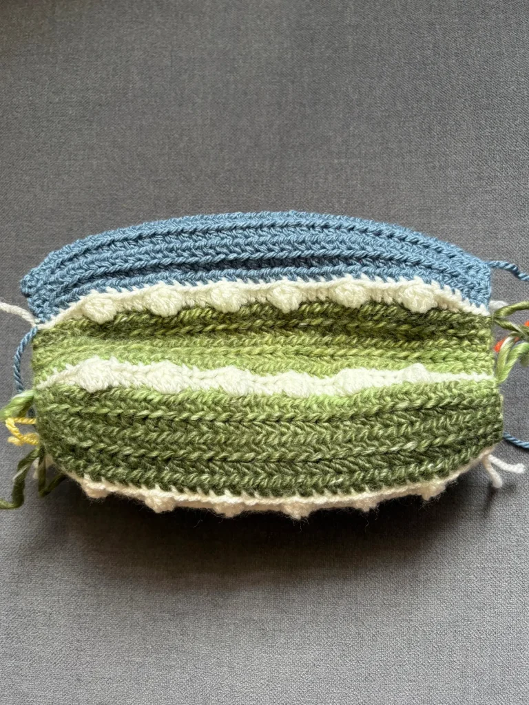 A crocheted oval swatch features alternating rows of blue, white, and green yarn, with textured and ridged patterns reminiscent of a charming pumpkin crochet pattern, laid flat on a gray surface.
