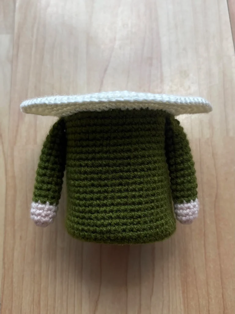 A crocheted green figure with two arms and a wide, flat white topโresembling a whimsical Toadstool Gnomeโis shown on a light wooden surface.