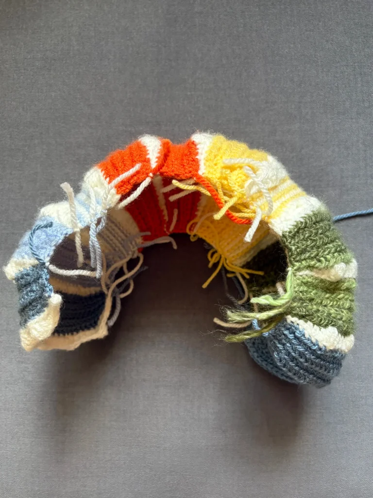 A partially finished knitted piece in rainbow stripes with loose yarn ends, displayed on a gray surface, sits beside a charming pumpkin crochet patternโperfect inspiration for anyone looking to try a crochet pumpkin pattern free this season.