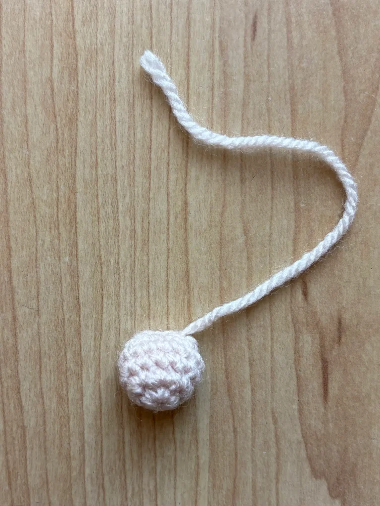 A small crocheted pink ball, reminiscent of a toadstool gnomeโs playful charm, with an attached yarn tail rests on a light wood surface.