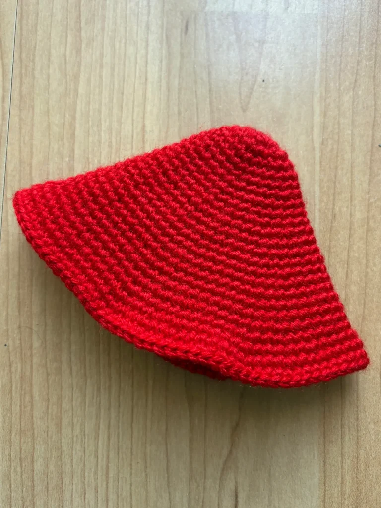 A red crochet object with a triangular shape, reminiscent of a Toadstool Gnomeโs hat, rests on a light wood surface.