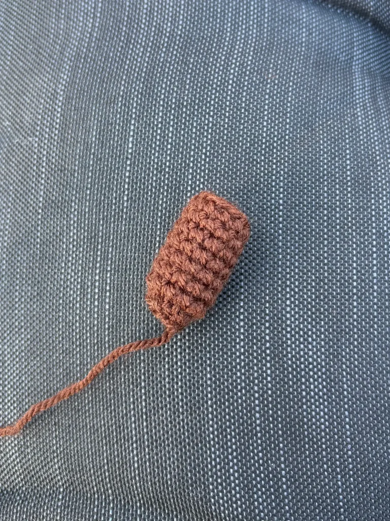 A small, brown crocheted cylinder made using a pumpkin crochet pattern, with a loose yarn tail, rests on a gray, textured fabric surface.