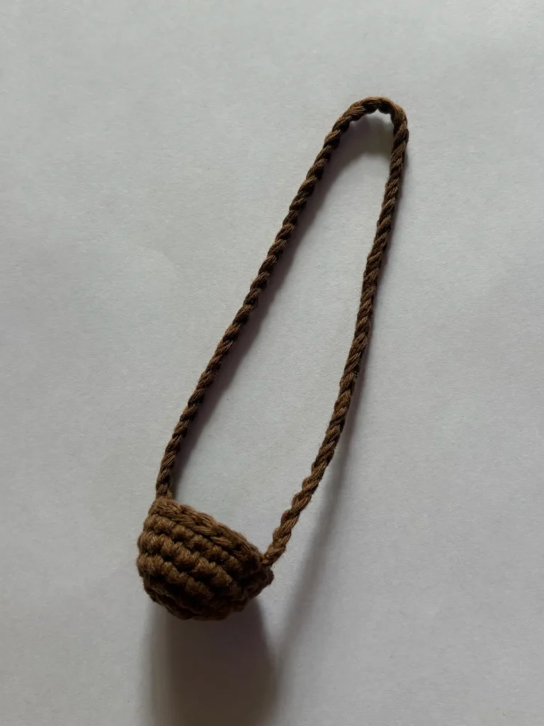 Small, brown, hand-crocheted basket with a long braided strap, placed on a plain light background.