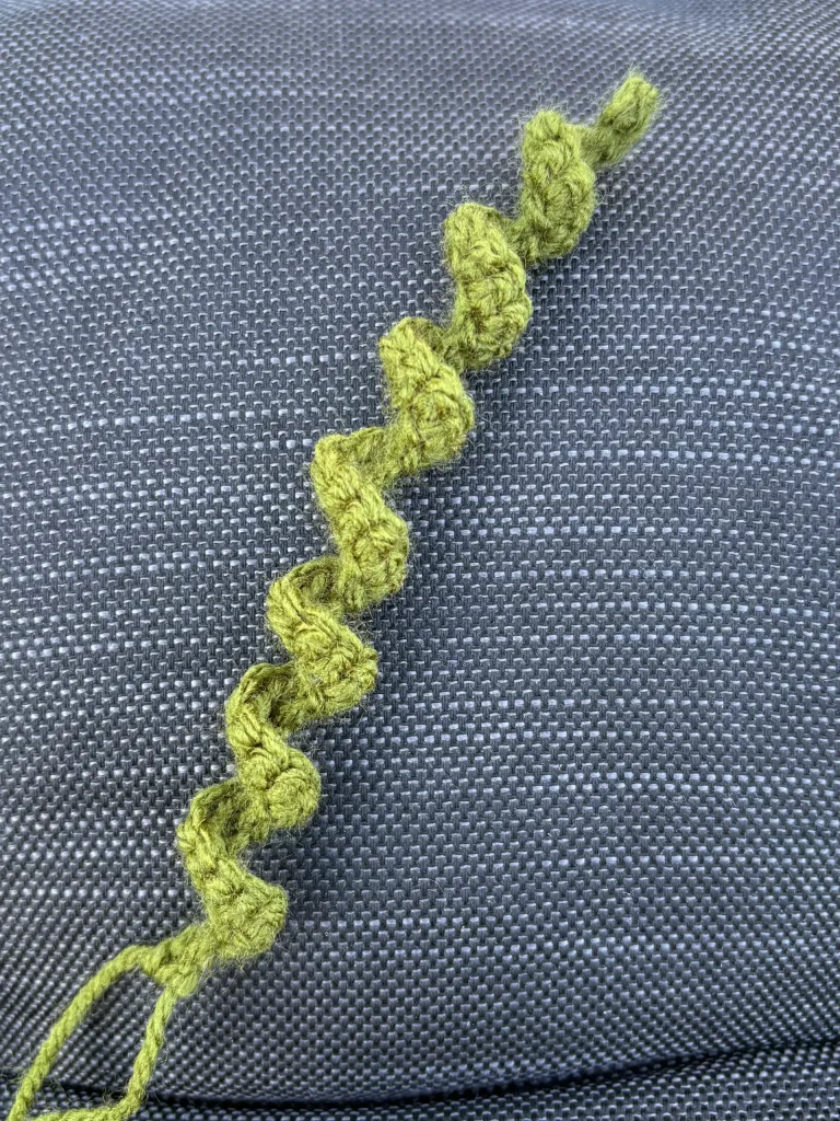 A green crocheted spiral, reminiscent of a free crochet pumpkin pattern, lies on a textured dark gray fabric surface.