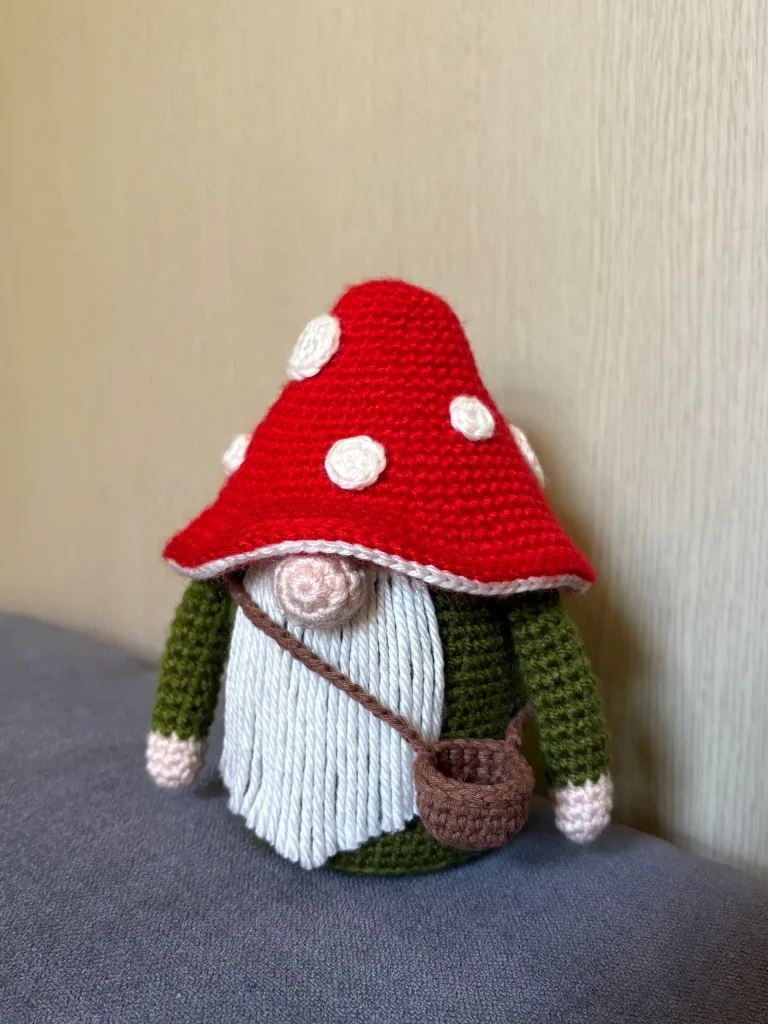 A crochet gnome figurine with a red mushroom cap hat, green sleeves, a white beard, and a small brown satchel is placed against a beige wallโmaking it an adorable companion for autumn dรฉcor or to display alongside your favorite crochet pumpkin pattern.
