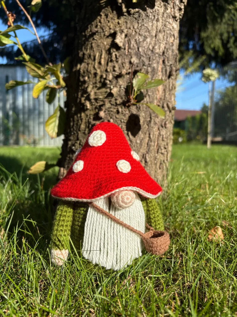 A crochet gnome doll with a red mushroom cap hat and green outfit sits on grass leaning against a tree trunk in a sunny outdoor setting, perfect for inspiring your next projectโwhether itโs this whimsical scene or trying out a free crochet pumpkin pattern to add even more charm.