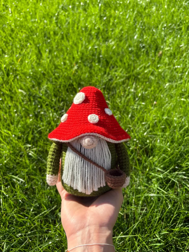 A hand holds a crocheted Toadstool Gnome doll with a red mushroom hat and white spots, standing on bright green grass in sunlight.