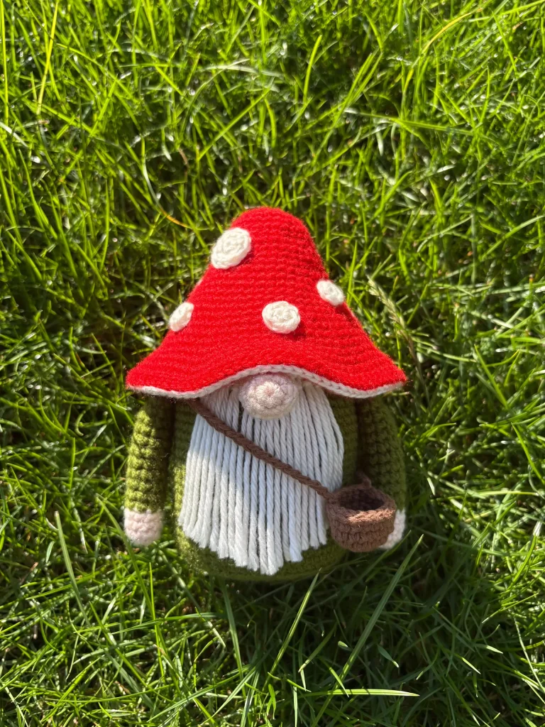 A crocheted Toadstool Gnome with a red mushroom cap hat and a white beard stands on green grass, holding a small brown bagโan enchanting companion for your next free crochet pumpkin pattern project.