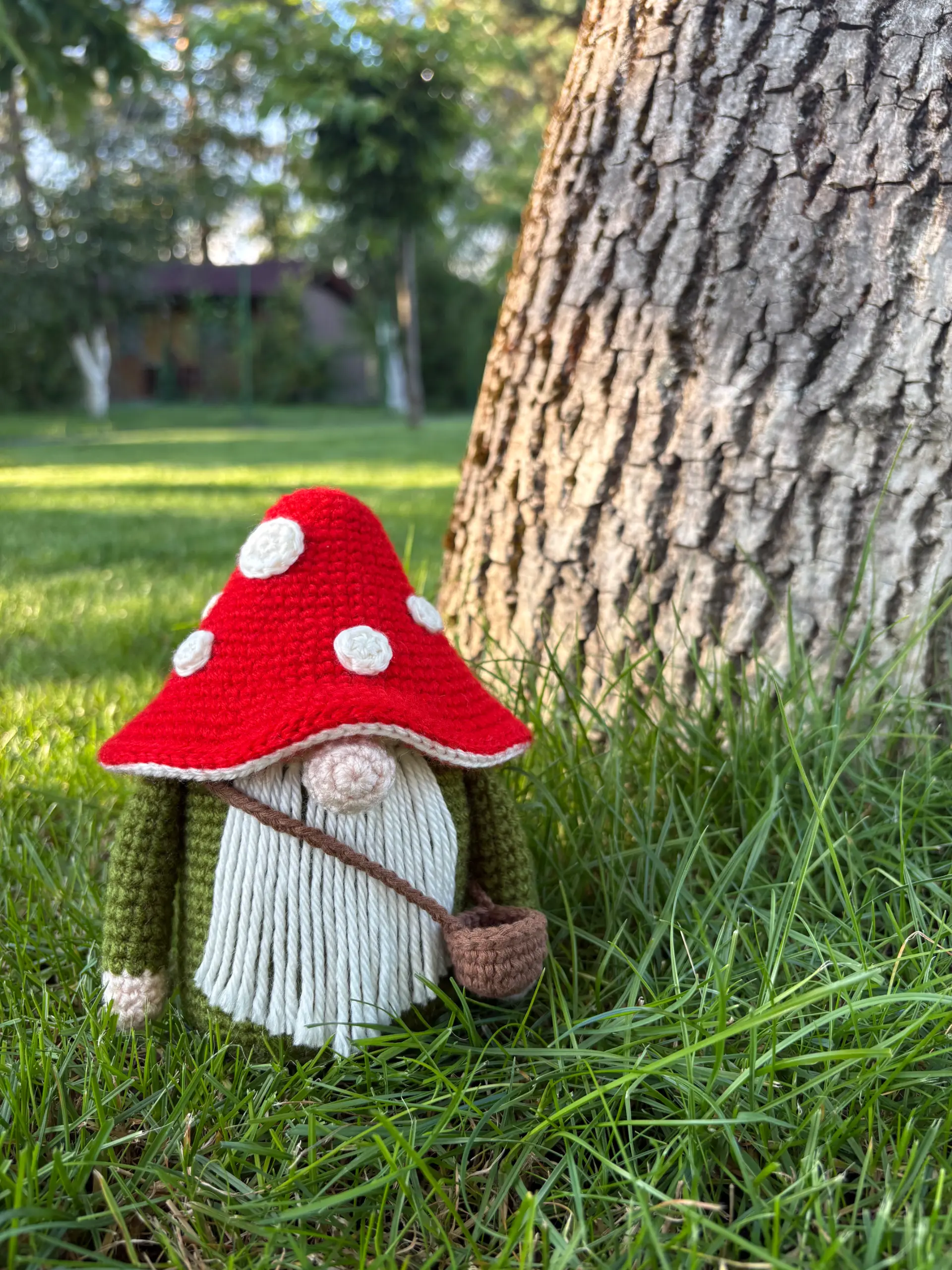 A crochet gnome figure with a red and white mushroom cap hat and a small bag stands in grass beside a tree trunk, ready to gather supplies for his next crochet pumpkin pattern.