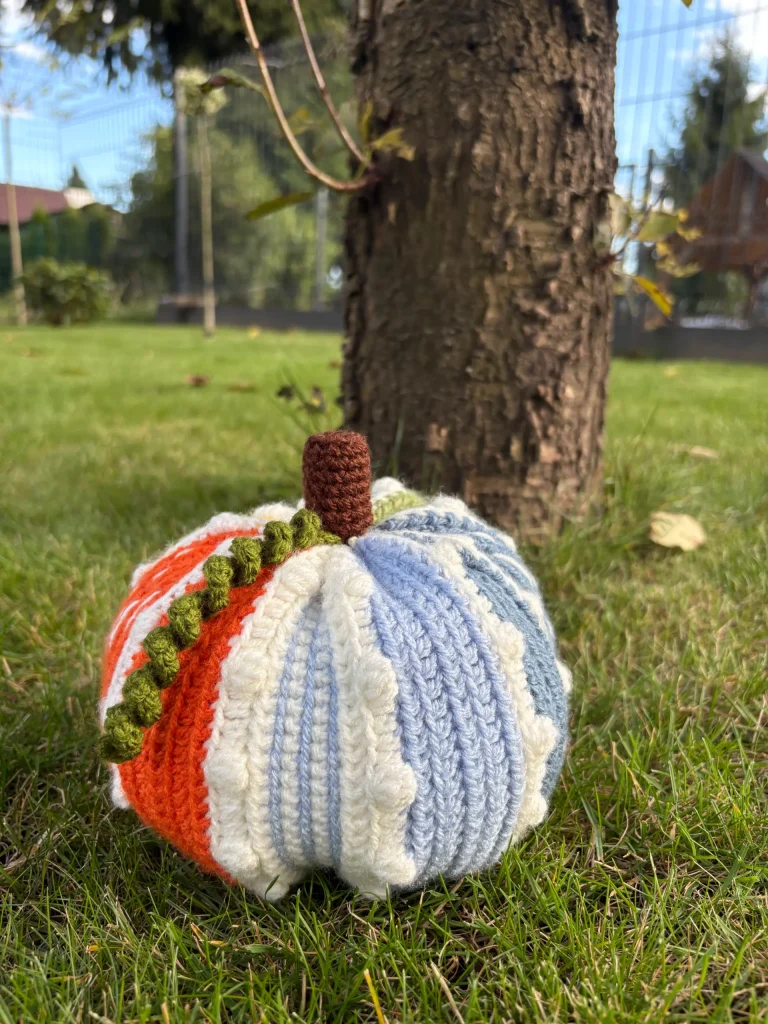 A crochet multicolored pumpkin with a brown stem sits on green grass in front of a tree trunk outdoors, making it the perfect inspiration for anyone searching for a free crochet pumpkin pattern.