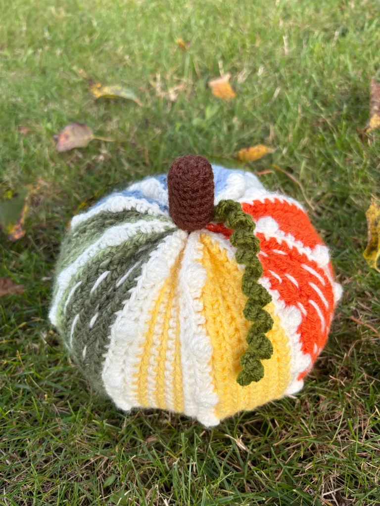 A multicolored crocheted pumpkin with a brown stem and green vine sits on grass, showcasing a charming design inspired by a free crochet pumpkin pattern.