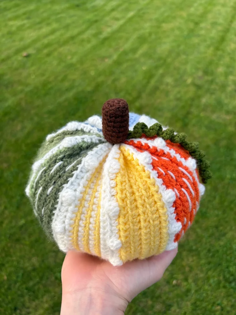 A hand holds a multicolored, crochet pumpkin, crafted from a free crochet pumpkin pattern, with green, yellow, white, and orange sections, a brown stem, and a green leaf. A grassy lawn is visible in the background.