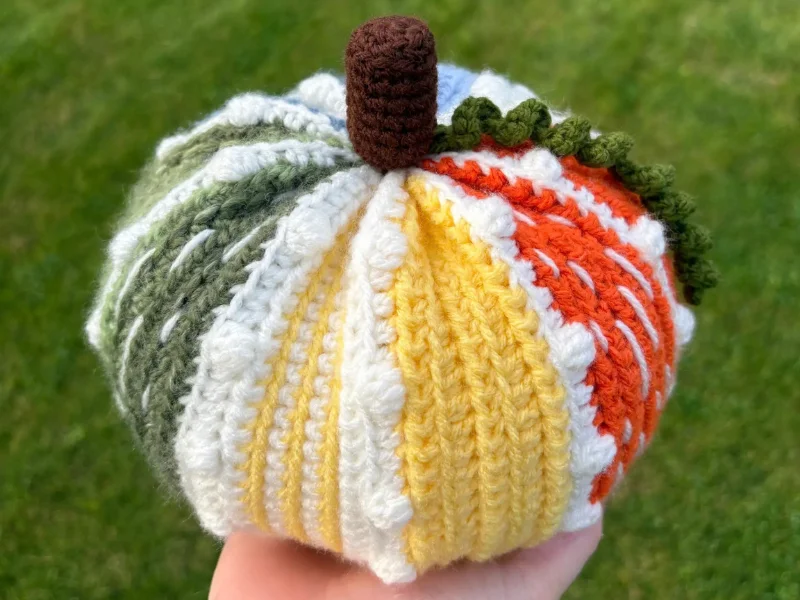 A hand holds a crochet pumpkin with vertical stripes in green, white, yellow, and orange yarn, featuring a brown stem and green detail, against a grassy background—perfect inspiration for your next crochet pumpkin pattern.