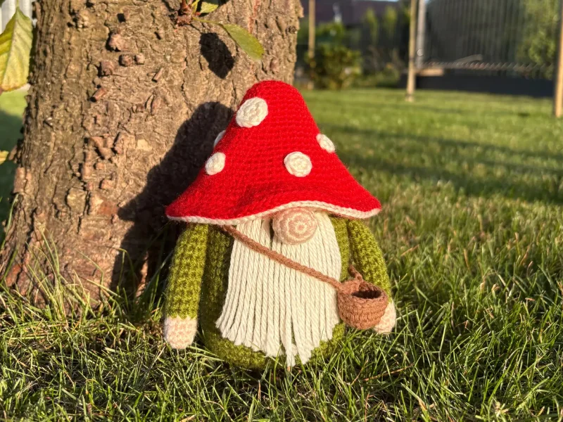 A crochet Toadstool Gnome with a red, white-spotted mushroom hat and a small brown bag stands against a tree on green grass in sunlight—perfect inspiration for your next crochet pumpkin pattern free project.