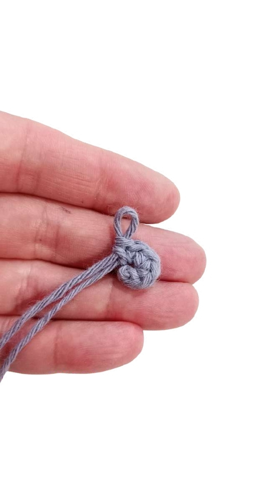 A hand holds a small gray crochet pattern piece shaped as a circle with a loop of yarn protruding from the top.