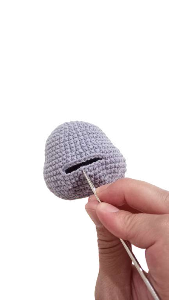 A hand holding a crochet hook inserts it into a slot on a round, grey piece for this pumpkin crochet pattern.