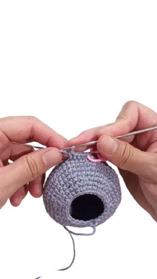 Hands crocheting a small, round grey object into an elephant amigurumi using a silver crochet hook and a pink stitch marker.
