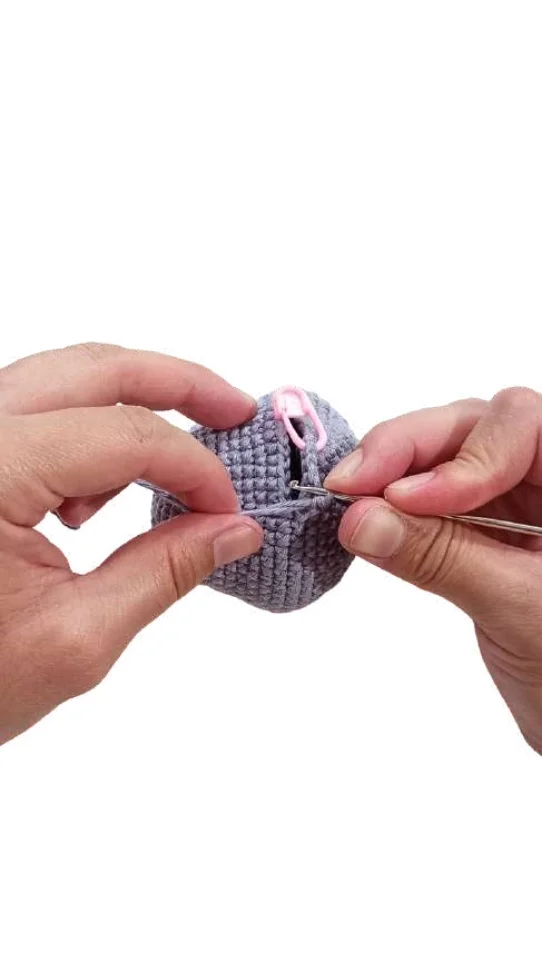 Hands using a crochet hook to work on a grey crocheted sphere, a step in this pumpkin crochet pattern, marked with a pink stitch marker.