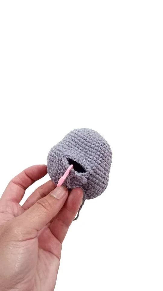 A hand holding a small, gray, unfinished piece from a crochet pumpkin pattern, accented with a pink stitch marker.