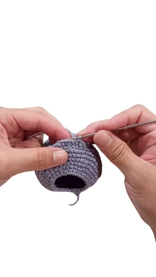 Hands holding a metal crochet hook while working on a grey crocheted sphere for an elephant amigurumi pattern.