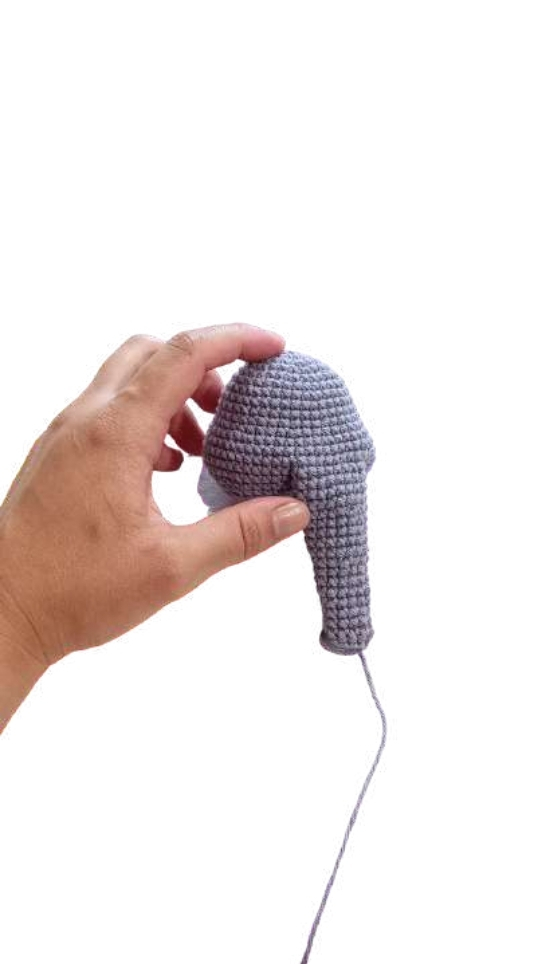 A hand holding a grey crocheted piece, reminiscent of a crochet pumpkin pattern, shaped like an elephant’s head and trunk.