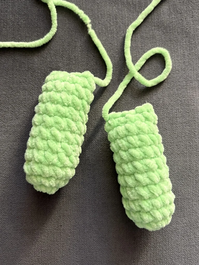 Two light green, crocheted Frankenstein plushie cylinders with yarn tails on a gray surface.