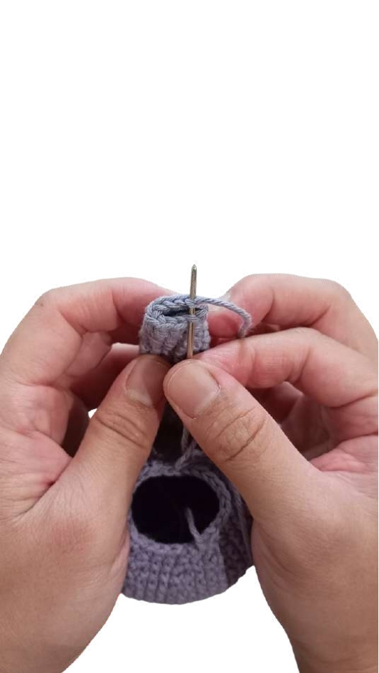Hands stitching the top edge of a crocheted lavender pumpkin pattern with a yarn needle.