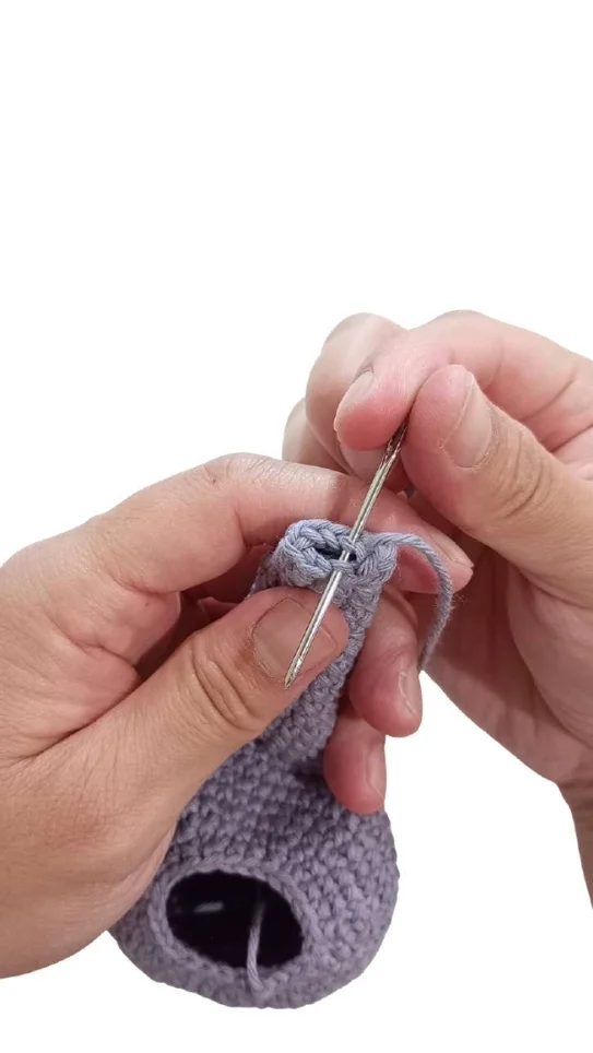 Hands using a metal needle to sew closed the top of a purple pumpkin crochet pattern.